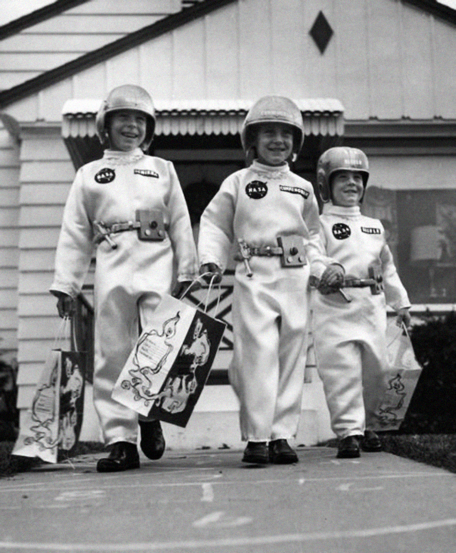Three children dressed as astronauts with helmets and matching suits walk on a sidewalk, smiling and holding Halloween treat bags in front of a house.