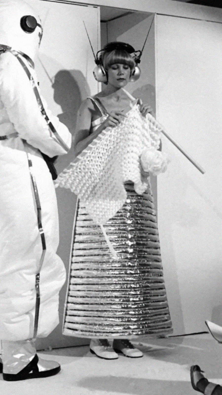 A woman in a metallic, futuristic dress and headphones knits with oversized needles, while a person in a white spacesuit with a helmet and antennae stands nearby. The scene appears science fiction-themed.