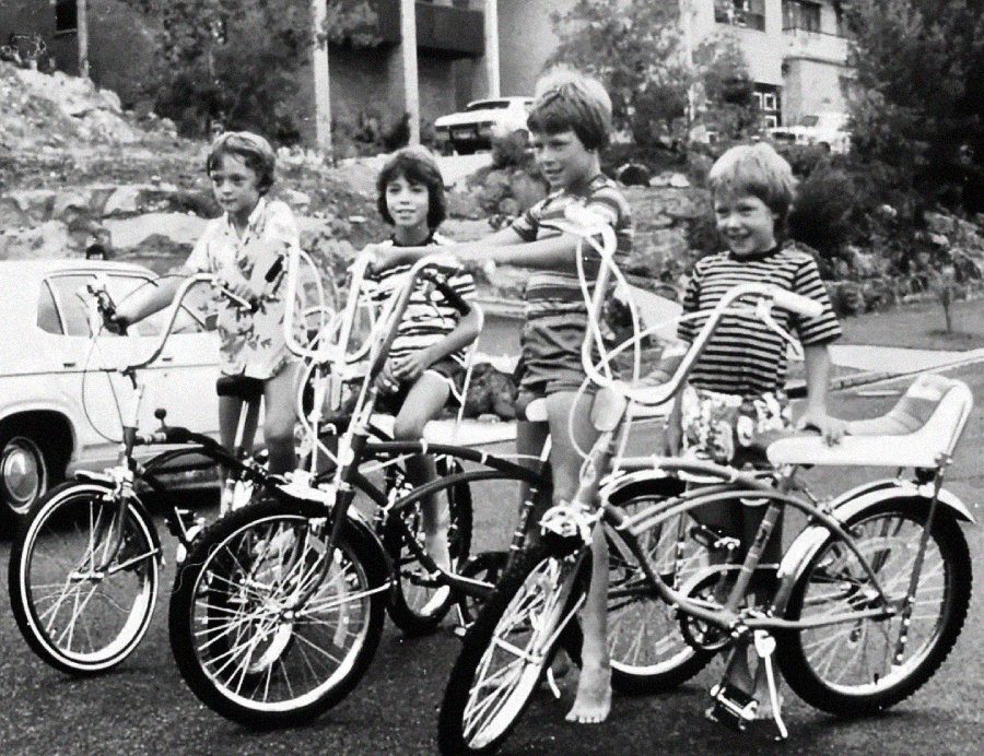 Four children pose outdoors with vintage bicycles, smiling. The scene appears to be from past decades, with cars and houses visible in the background. Some children are barefoot and all are dressed in casual clothes.