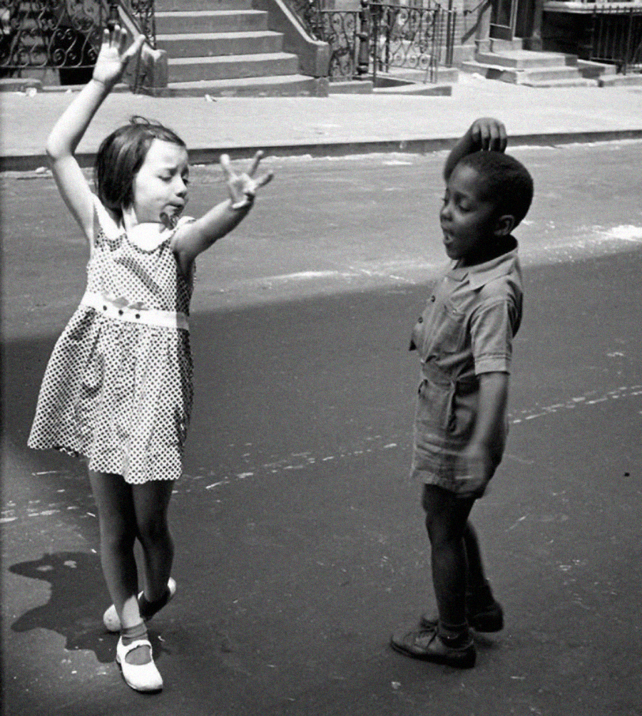 A young girl and boy dance joyfully together on a city street. The girl wears a polka dot dress, and the boy wears a collared shirt and shorts. Their arms are raised playfully, and they smile at each other.