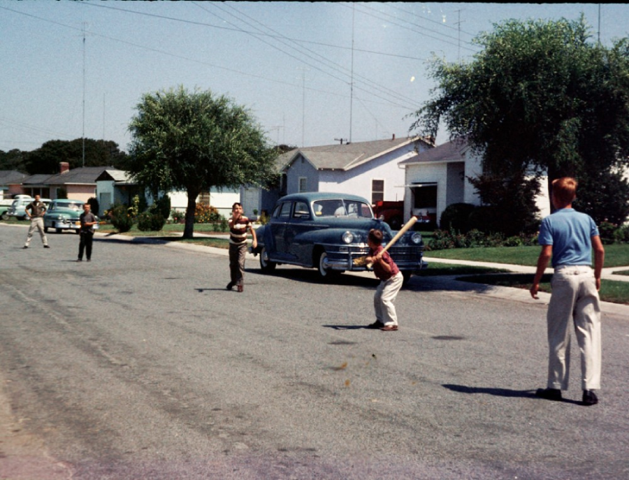Four boys play baseball on a quiet suburban street lined with vintage cars and houses. One boy is batting while others field. The scene looks like it's from the mid-20th century. Trees and lawns border the road.