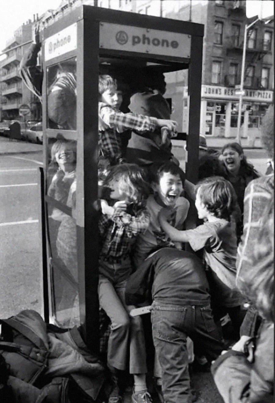 A group of children laughs and squeezes together inside an old phone booth on a city street, some climbing over each other, capturing a playful and chaotic scene.