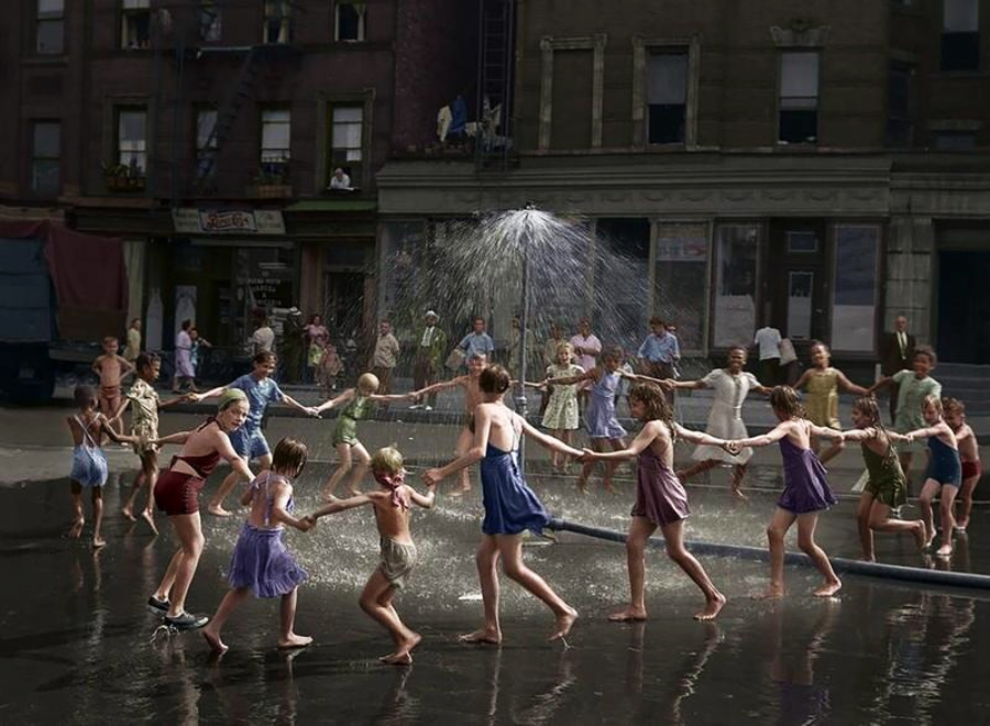 Children in swimsuits and summer clothes hold hands, forming a circle around a spray of water from a fire hydrant in a city street, playing and laughing on a sunny day, with apartments and onlookers in the background.