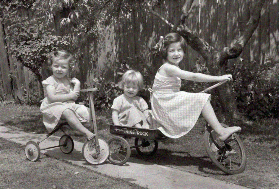 Three young children in dresses play outside. One rides a tricycle pulling a wagon with a child inside, while another sits on a small tricycle. Trees and a wooden fence are in the background. The photo is black and white.