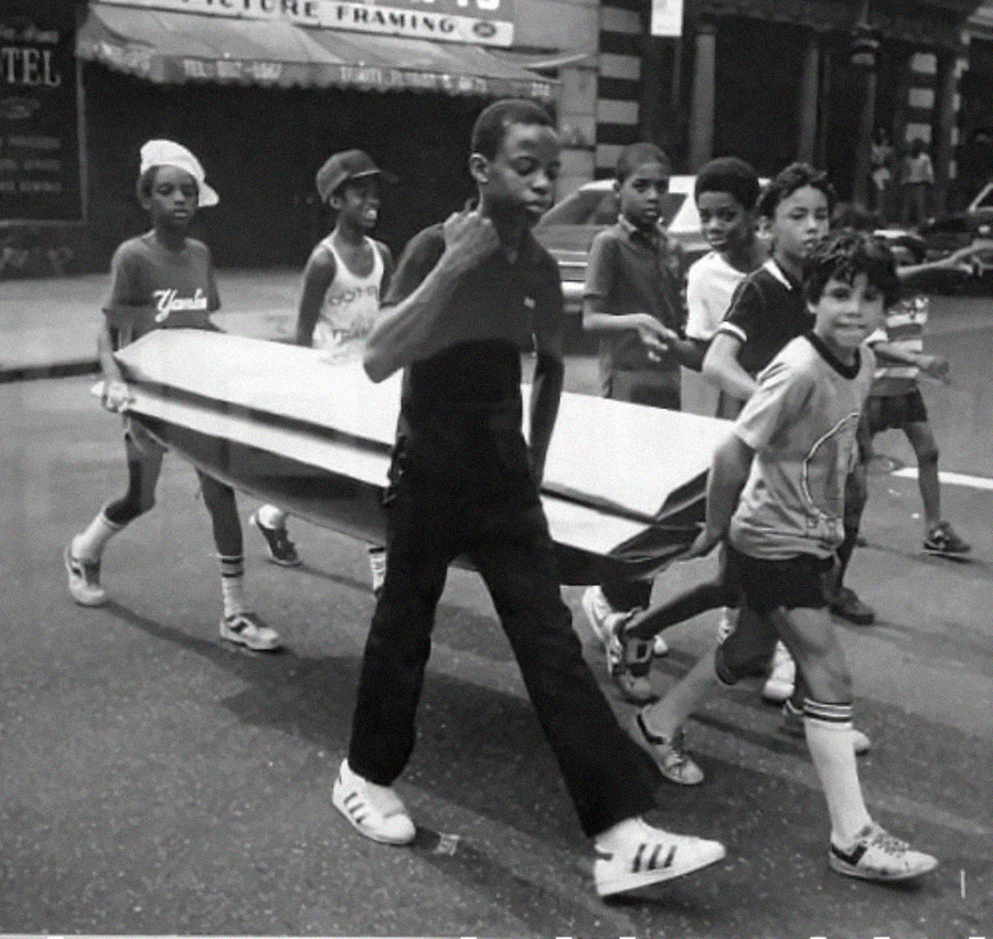 A group of children walk down a city street carrying a large rectangular object together, appearing cheerful and energetic. Buildings and parked cars are visible in the background.