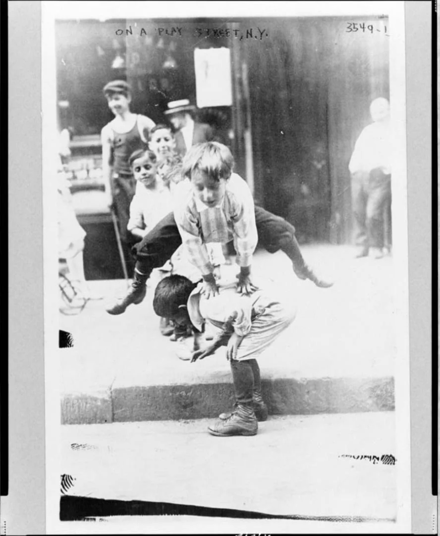 Black-and-white photo of children playing leapfrog on a sidewalk, with one boy leaping over another who is bent over. Several other kids and an adult watch and smile in the background. The setting appears to be an urban street.