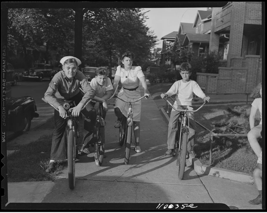 Four kids are lined up with their bicycles on a residential sidewalk, looking toward the camera. Trees and houses are visible in the background on a sunny day. One child is dressed as a sailor.