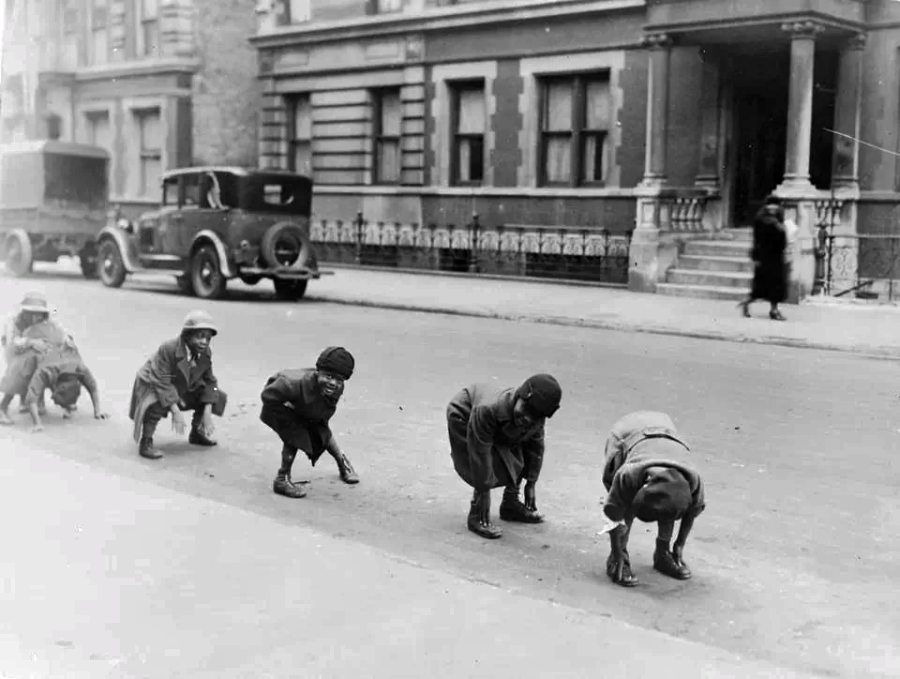 Five children play leapfrog in a line on a city street, wearing coats and hats, with vintage cars and buildings in the background; a blurred figure walks on the sidewalk.