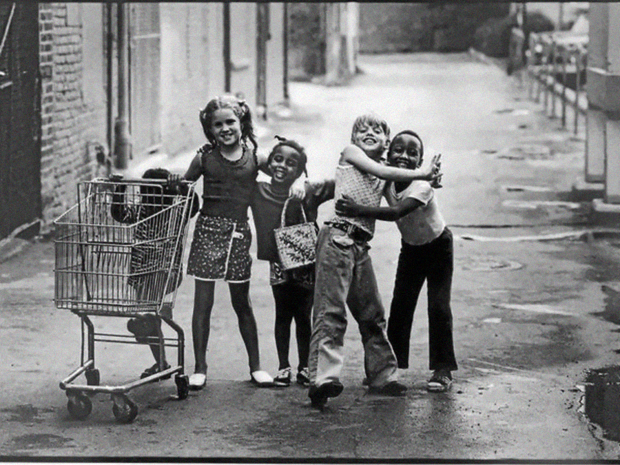 Four children stand together in an alley, smiling and hugging. One child hugs another while two others stand close beside them. A shopping cart is on the left side. The mood is joyful and playful.