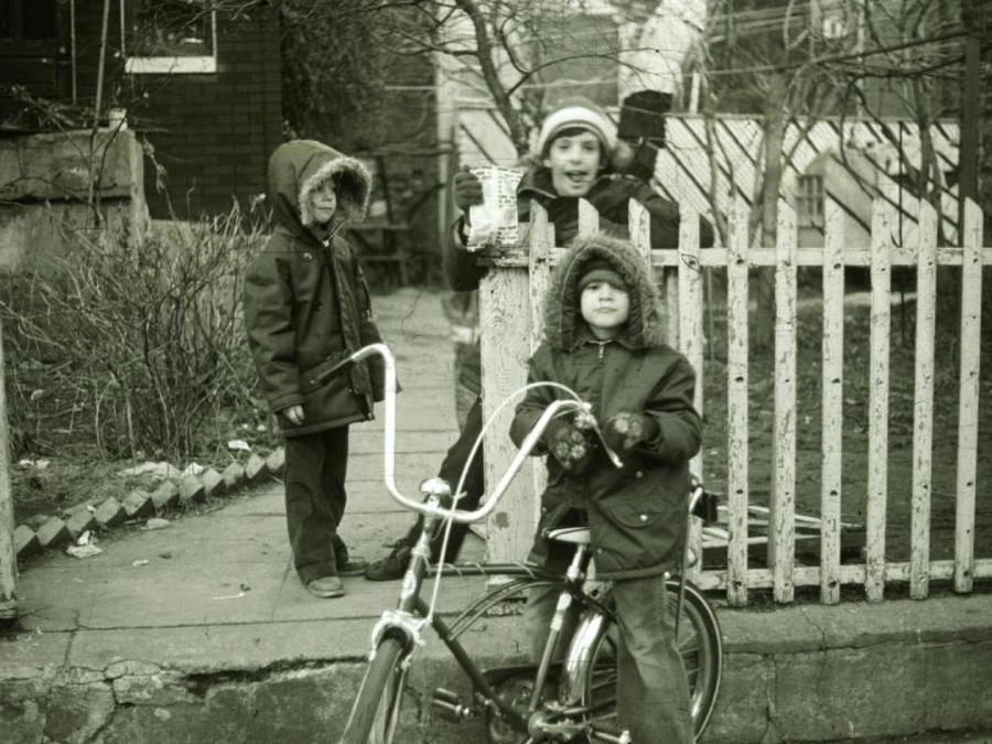 Three children in winter coats stand outside near a picket fence; one sits on a bike, another stands beside, and the third holds up an object, all appearing cheerful on a cold day.