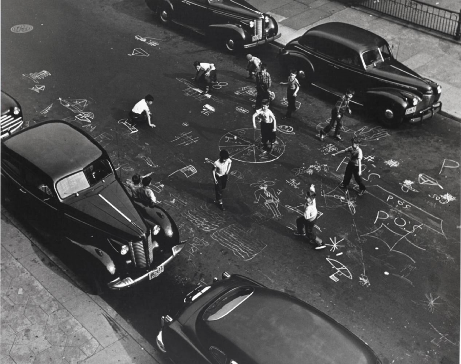 Children play on a city street covered in chalk drawings, surrounded by parked vintage cars. The scene is lively, viewed from above, with kids interacting and sketching on the pavement.