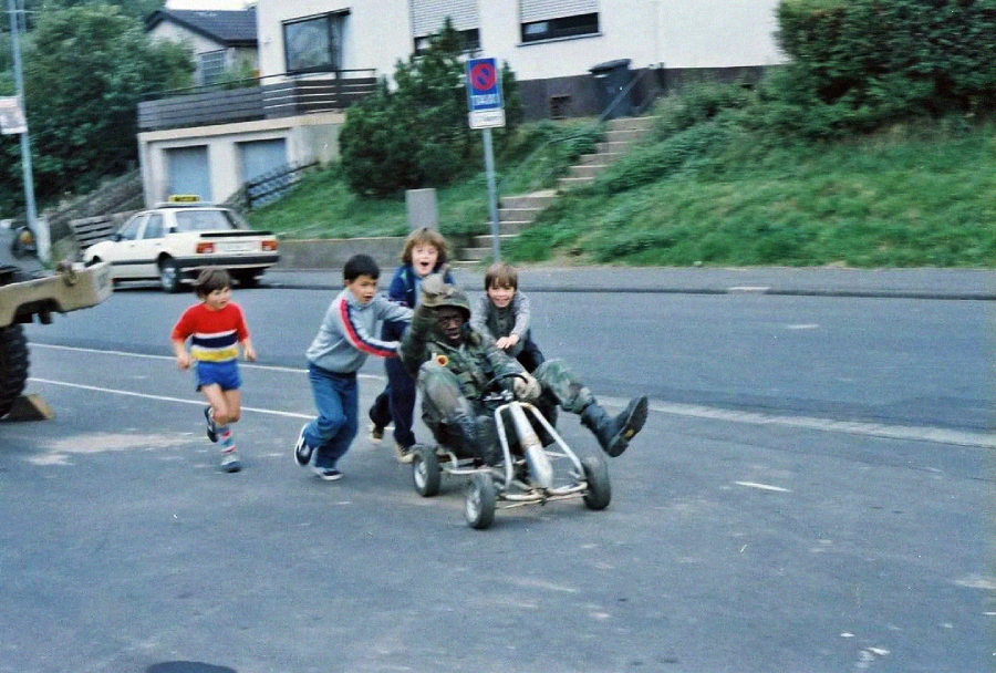 Four boys push a go-kart with a person dressed in camouflage gear riding in it, all smiling and running on a suburban street with houses and parked cars in the background.