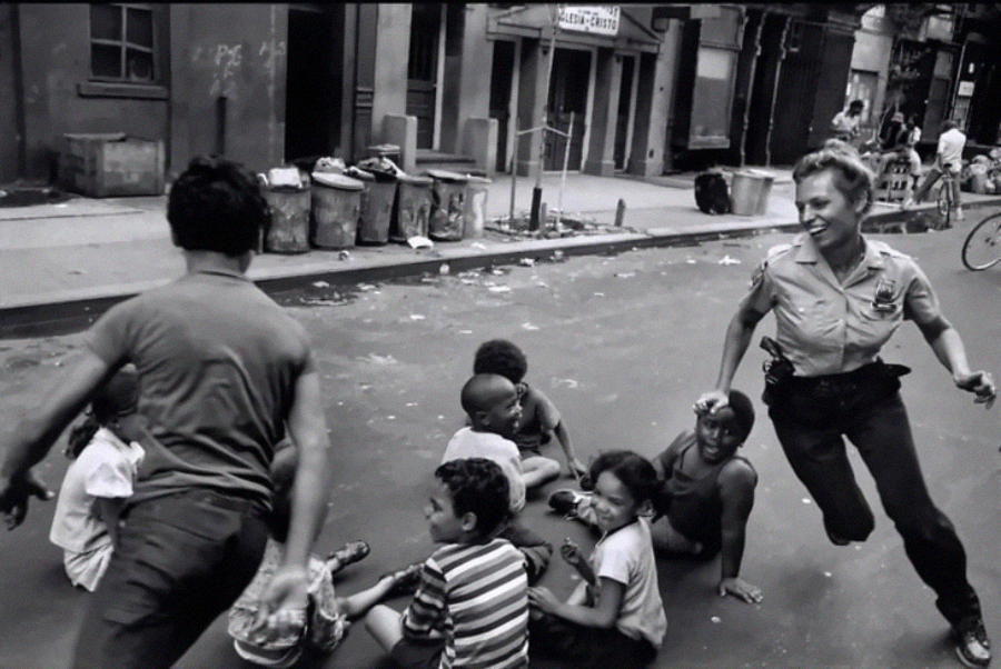 A police officer playfully chases a group of smiling children sitting and running in the middle of a city street lined with old buildings and trash cans.