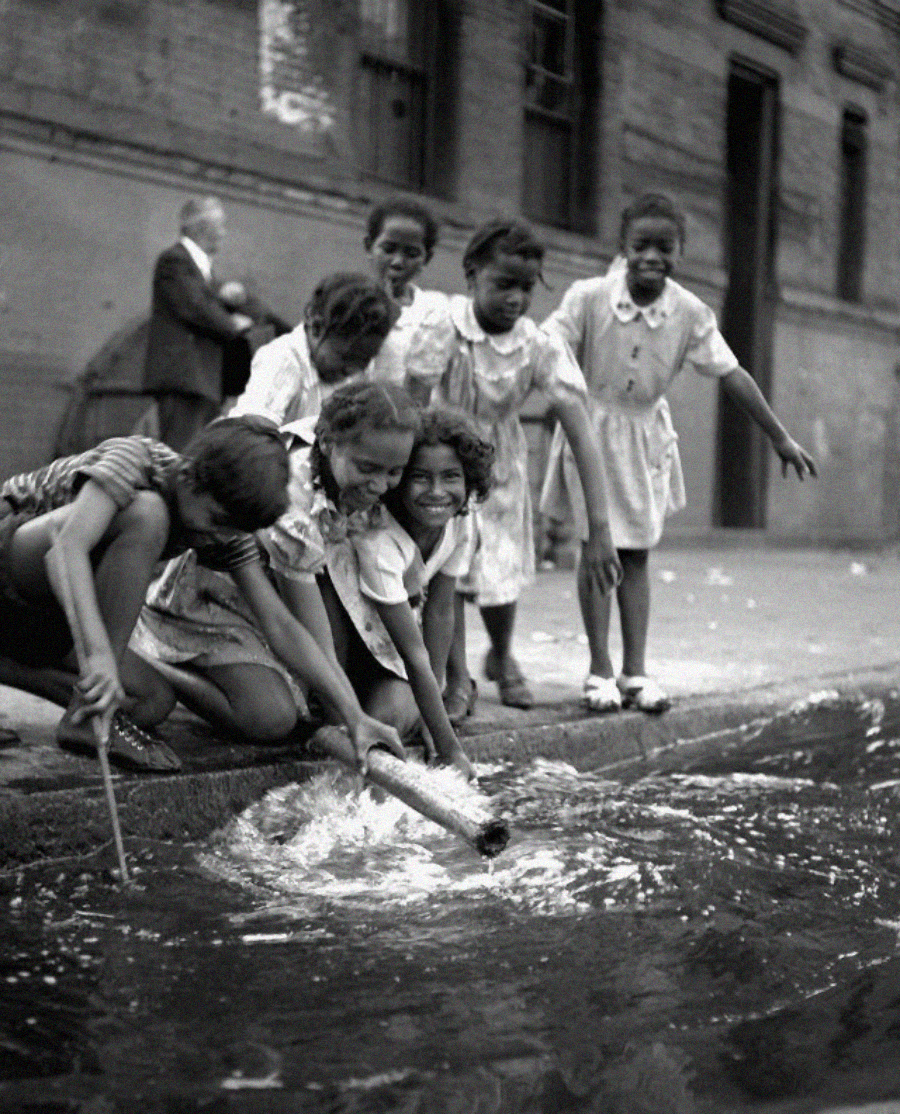 A group of six young girls, smiling and laughing, play together on a city street curb, splashing water with sticks in a puddle. An adult stands in the background near a brick building.