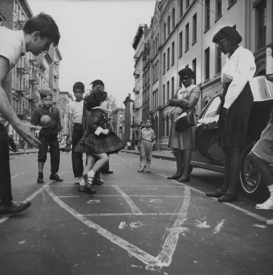 Children play hopscotch on a city street while adults watch nearby; buildings and parked cars line the background, creating a lively urban scene.