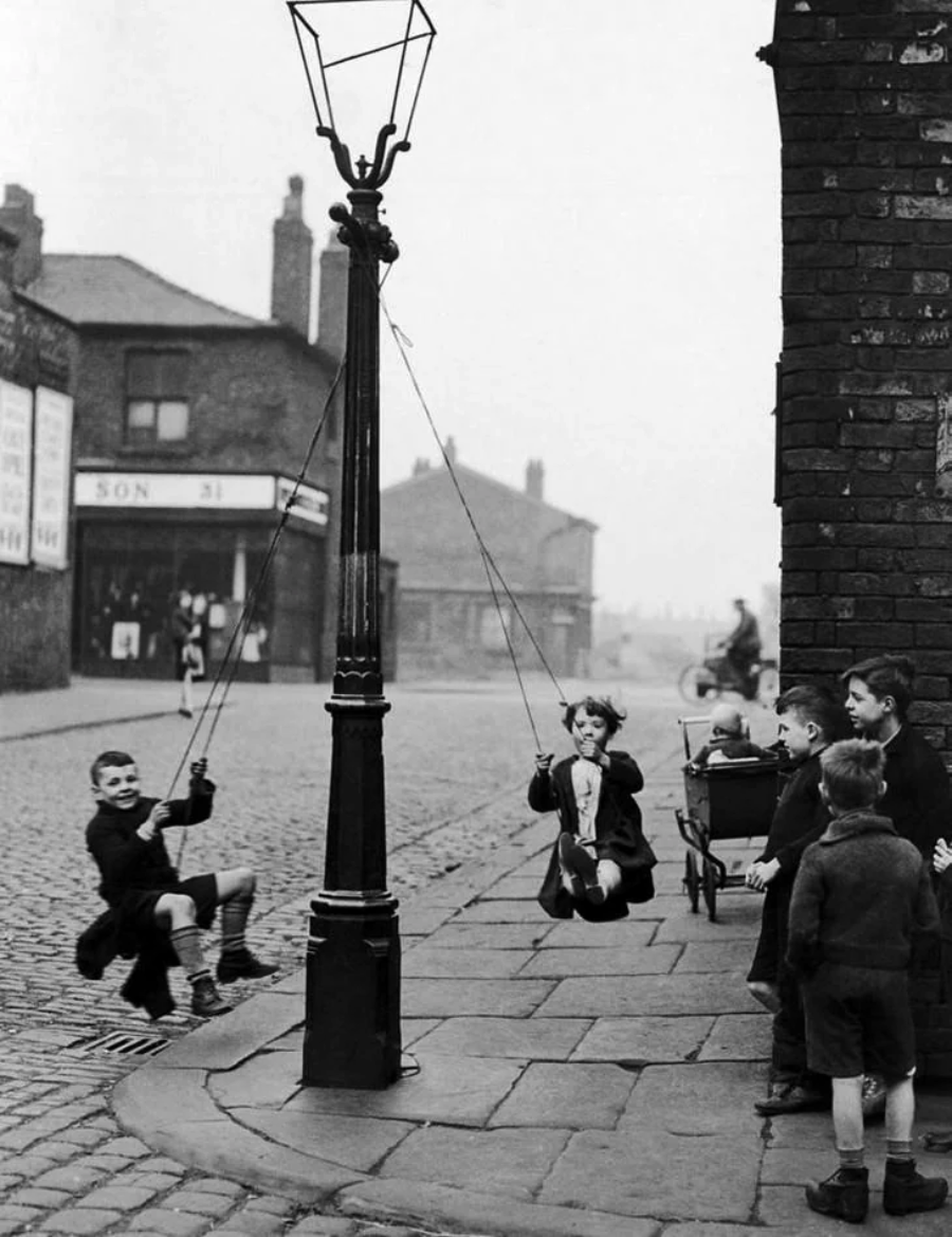 Black-and-white photo of children playing on swings attached to a streetlamp on a cobblestone street, while other children watch nearby; old buildings and a person on a bicycle are in the background.