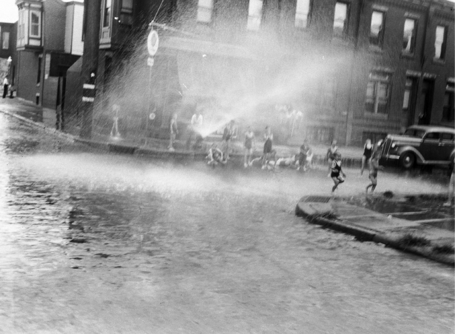 Children run and play in the spray of a fire hydrant on a cobblestone street in front of brick buildings, while others watch and sit on the sidewalk in the background.