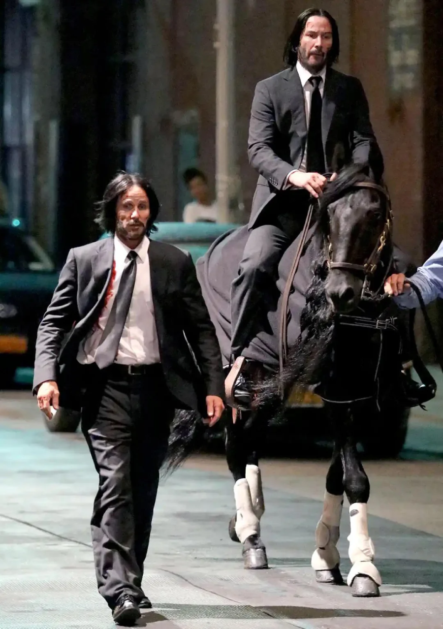 Two men in black suits, one walking with a blood-stained shirt and the other riding a black horse, move through a city street at night. Both have long dark hair and beards, and the horse wears white leg wraps.