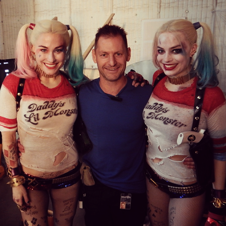 Three people pose for a photo; the two on the sides are dressed as Harley Quinn with "Daddy's Lil Monster" shirts, shorts, and pigtails, while the person in the middle wears a blue shirt and smiles.