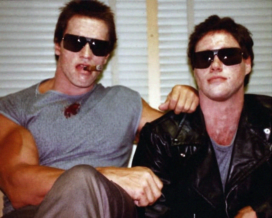 Two men with messy faces and sunglasses sit side by side; one wears a sleeveless shirt and smokes a cigar, while the other wears a black leather jacket. Both look relaxed against a backdrop of white blinds.
