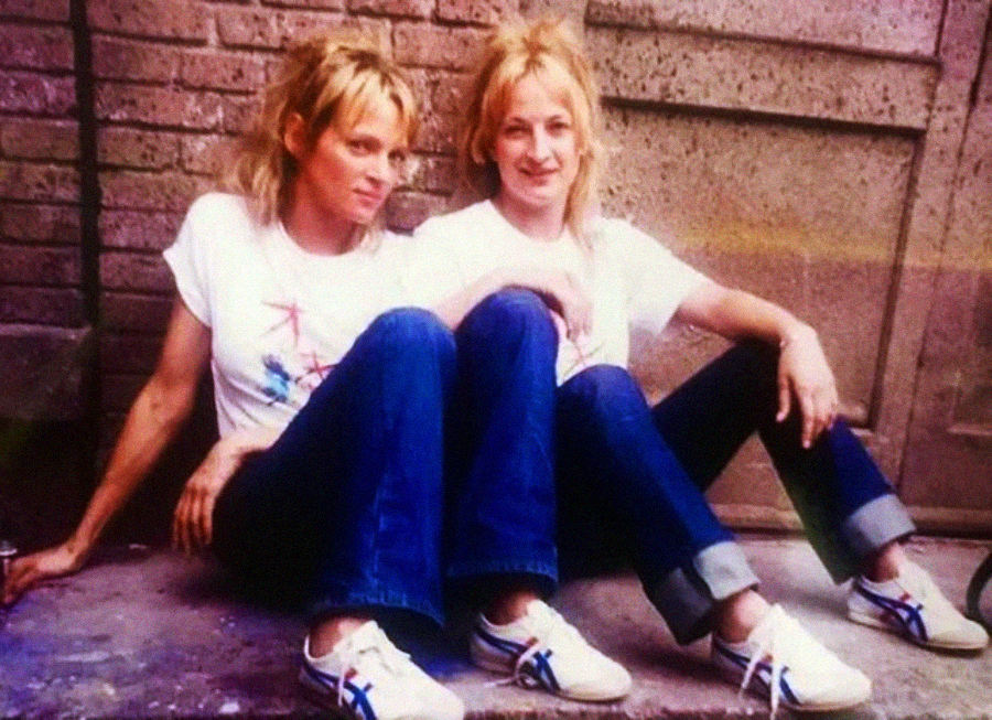 Two women with blonde hair, wearing matching white T-shirts, blue jeans, and white sneakers with blue stripes, sit on the ground against a brick wall and a concrete door, appearing relaxed and casual.