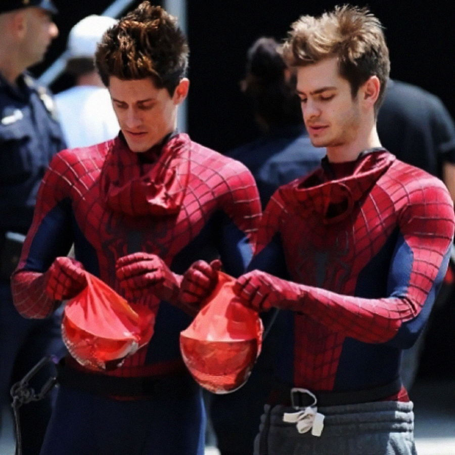 Two men dressed in Spider-Man costumes stand side by side, holding their red Spider-Man masks in their hands. They appear to be on a film set, with people in the background and focused expressions on their faces.