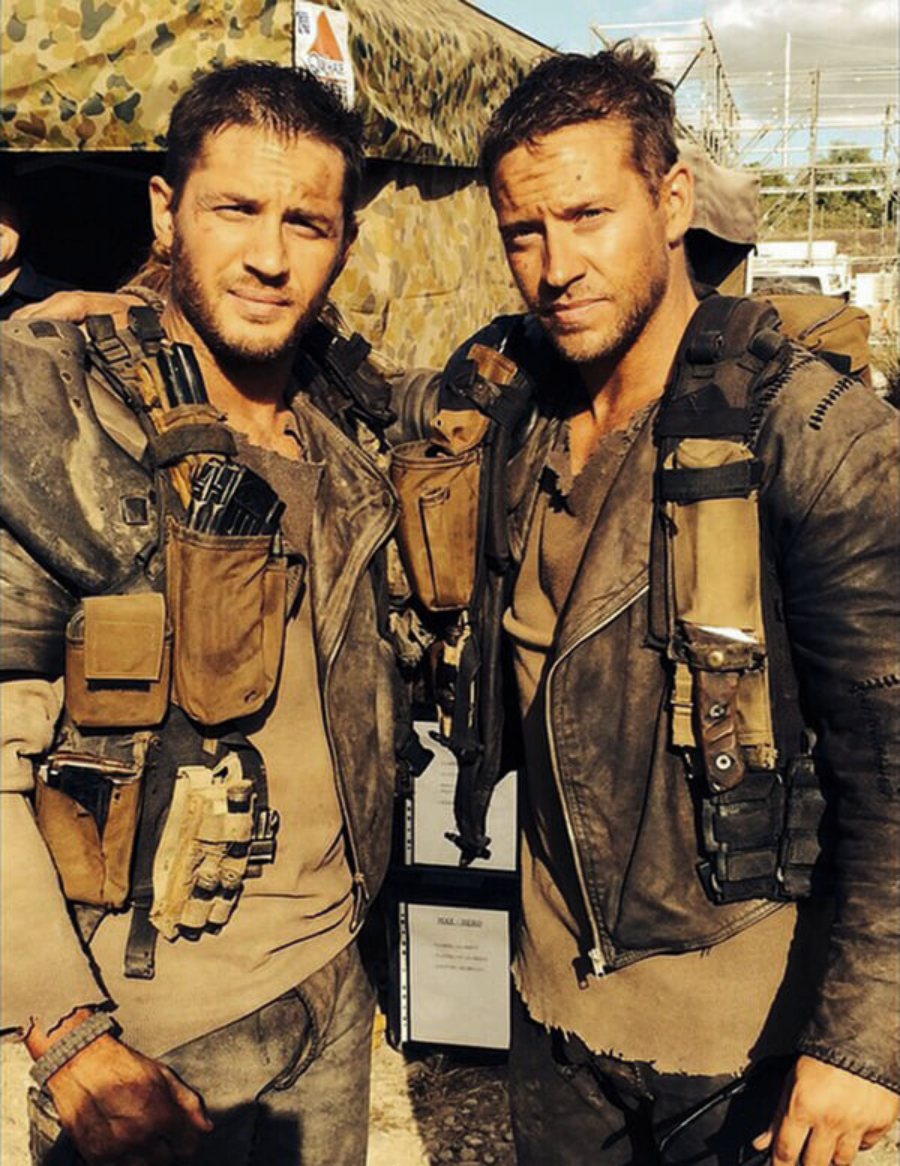 Two men in rugged, tactical outfits with brown gear and jackets stand close together, appearing dirty and weathered. They are outdoors near a camouflage tent, suggesting a post-apocalyptic or military setting.