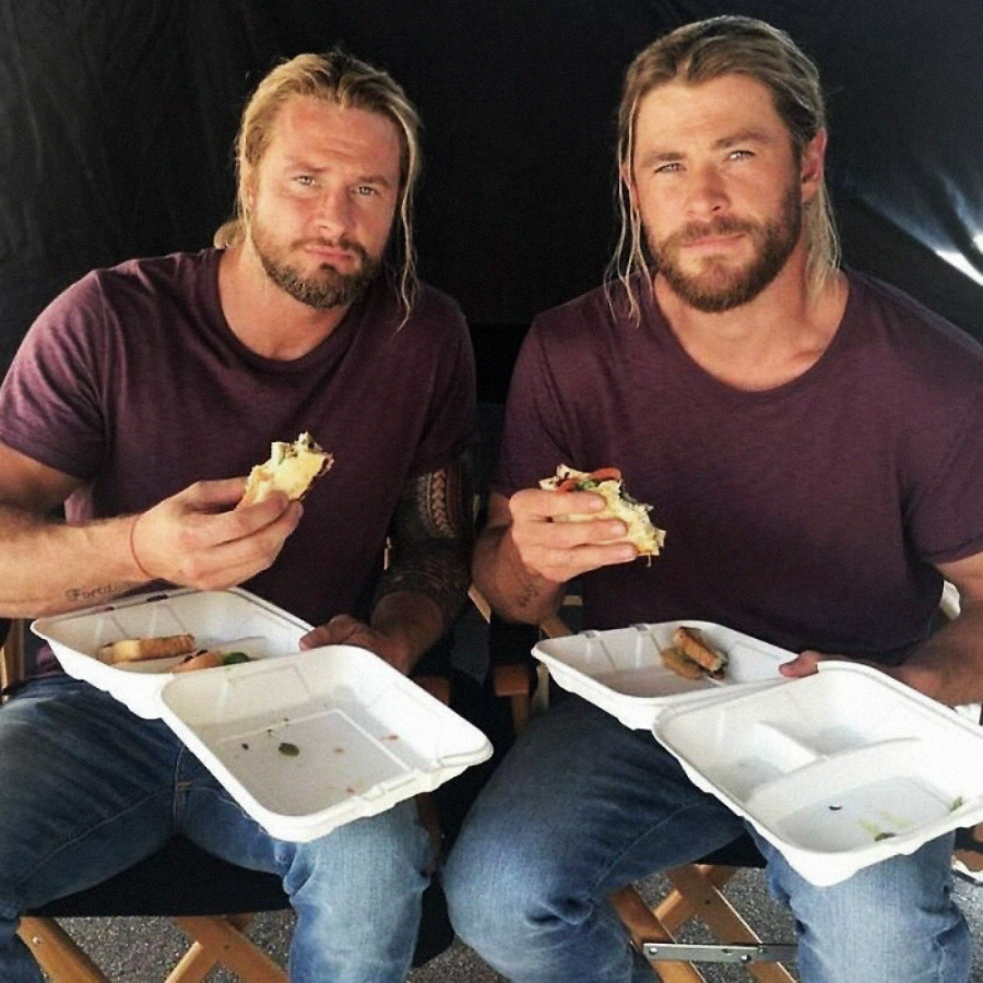 Two men with long blond hair and beards, wearing matching purple t-shirts and jeans, sit side by side on chairs eating sandwiches from white takeout containers. They look at the camera, appearing relaxed and casual.