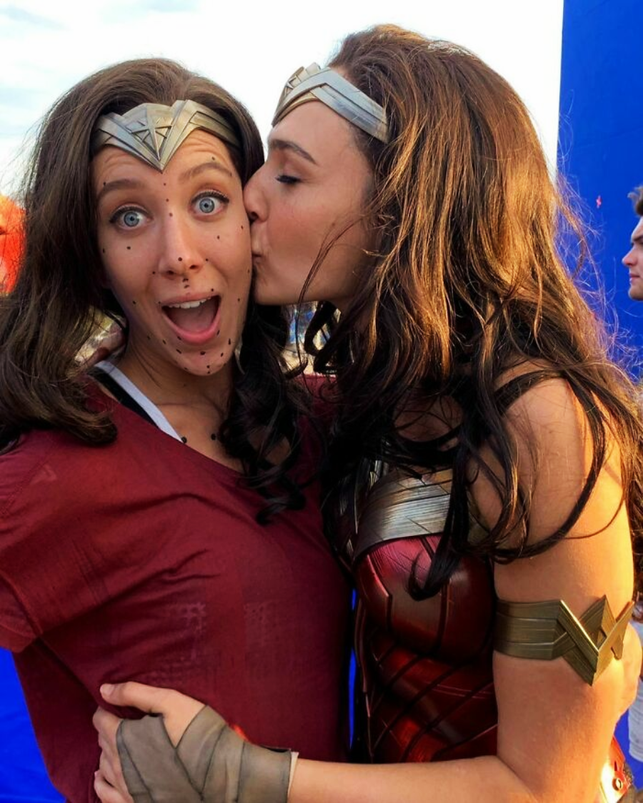 Two women dressed as Wonder Woman pose together. One kisses the other on the cheek; the other looks surprised and has facial markers for motion capture. Both are wearing brown wigs and Wonder Woman costumes.