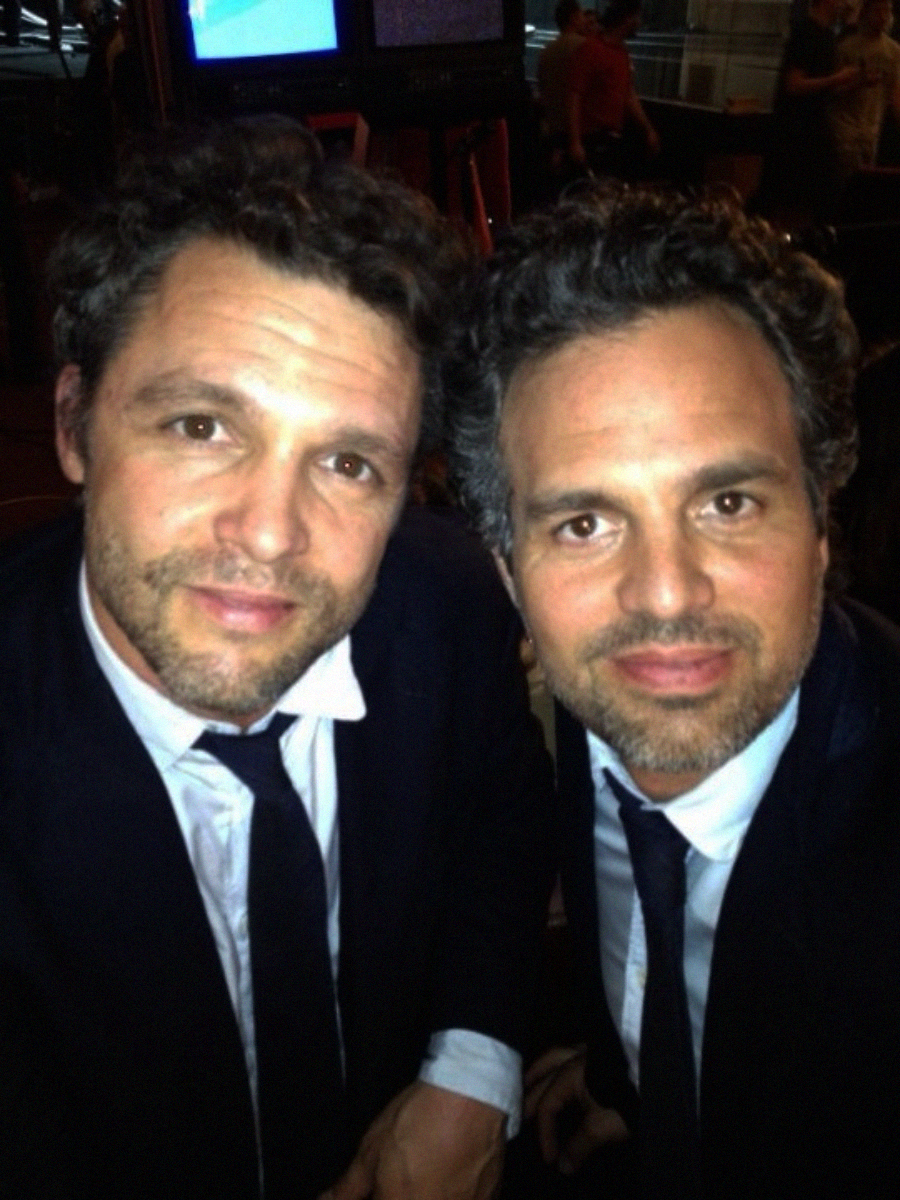 Two men with curly hair and beards, both wearing dark suits, white shirts, and black ties, sit closely together and look at the camera in a dimly lit indoor setting.