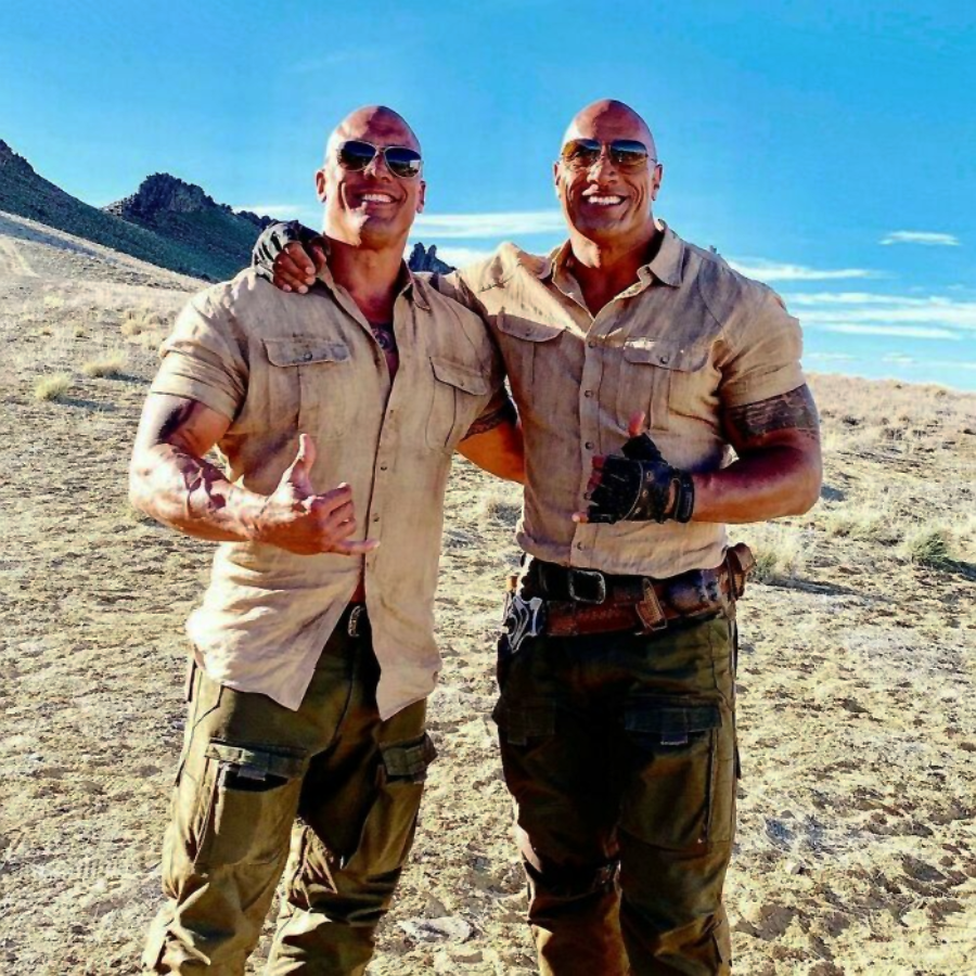 Two muscular men wearing sunglasses, short-sleeve khaki shirts, and cargo pants stand side by side in a desert landscape, smiling with their arms around each other and giving thumbs up.