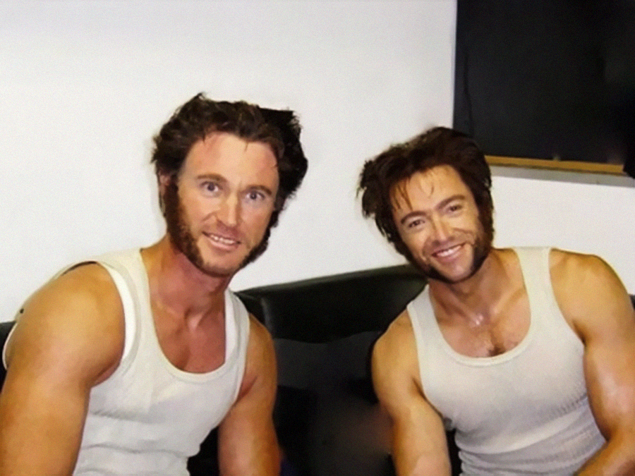 Two men wearing white tank tops and sporting similar sideburns and hairstyles sit side by side, smiling. They appear to be dressed as the same character, suggesting one may be a body double.