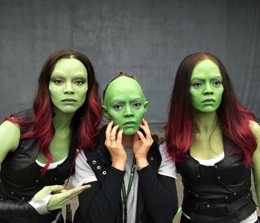 Three women pose together; two in black leather vests with green-painted skin and red-tipped hair, and one in the center wearing a bald cap and green face makeup, holding the sides of her face while the others gesture toward her.