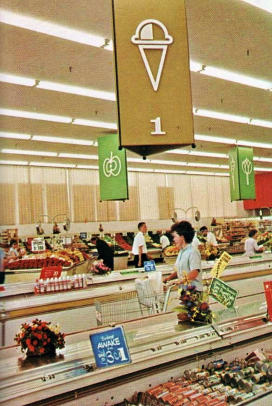 A woman pushes a shopping cart past frozen food aisles in a brightly lit, mid-century supermarket with signs overhead and shoppers in the background.