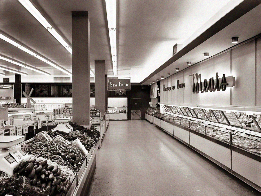 Black and white photo of a vintage supermarket interior showing produce on the left, a long refrigerated meat section on the right, and a sign above the back aisle reading “Sea Food.” The store appears clean and organized.