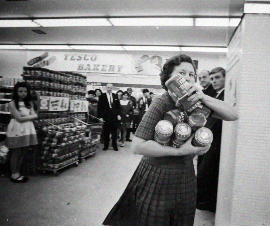 A woman clutches multiple cans while hurriedly walking in a busy Tesco supermarket, with onlookers and shelves of goods in the background.