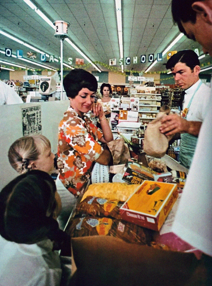 A woman with two children stands at a busy grocery store checkout counter filled with groceries as a cashier bags items. A "SCHOOL DAYS" banner hangs above in the store.