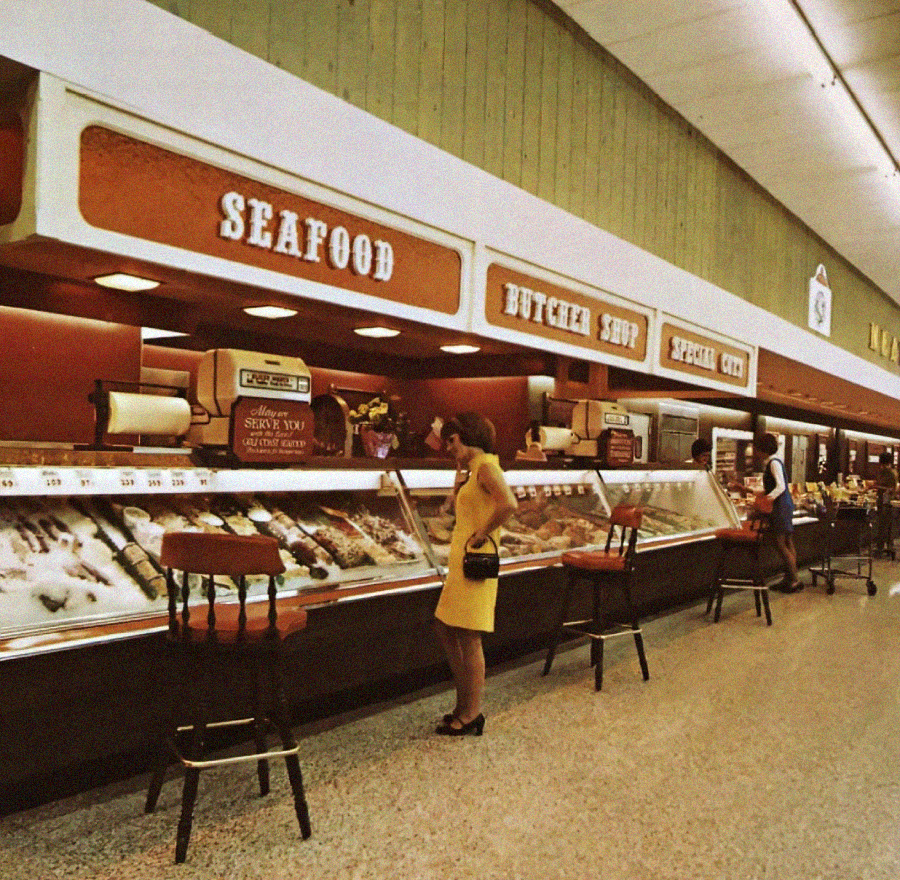 A woman in a yellow dress stands at a seafood counter in a vintage supermarket. There are signs above the counters reading "SEAFOOD," "BUTCHER SHOP," and "SPECIAL CUTS." Another person is visible in the background.