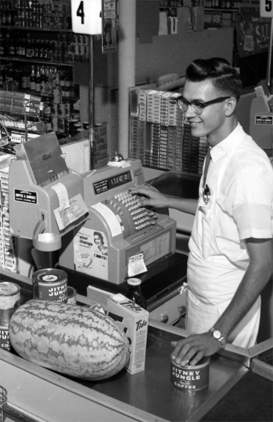 A young male cashier in a white uniform smiles while operating a vintage cash register in a grocery store; items on the counter include a watermelon, canned goods, and packaged food.
