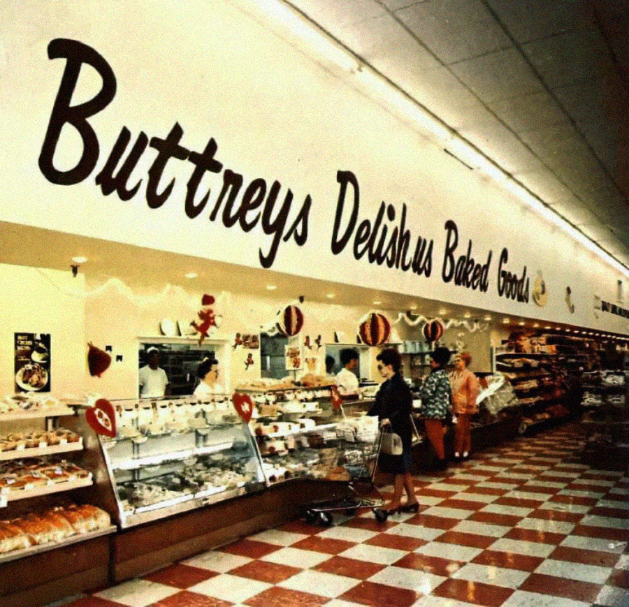 A vintage supermarket bakery section with a sign reading “Buttreys Delishus Baked Goods.” Shoppers browse displays of breads and pastries under warm lighting on a red-and-white checkered floor.