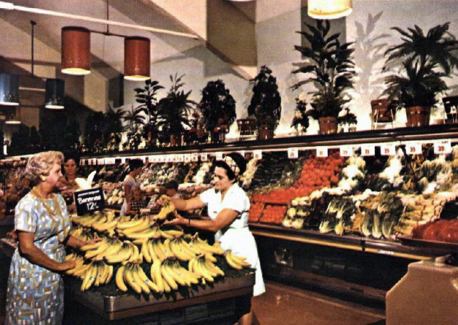 Two women in a vintage supermarket stand by a large banana display, surrounded by various fruits and vegetables, with potted plants on top of the produce shelves in the background.