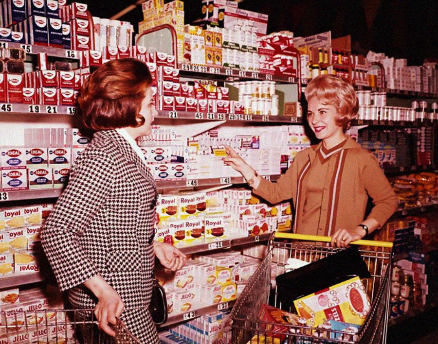 Two women in vintage clothing smile and chat while shopping in a brightly lit grocery store aisle, with one pushing a shopping cart filled with groceries and various products visible on the shelves behind them.
