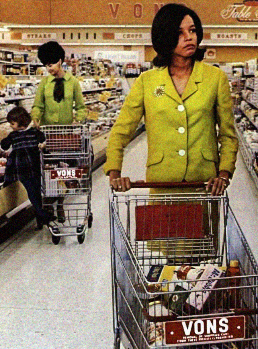 Two women in bright green outfits push shopping carts at a Vons supermarket. One woman stands in the foreground, while the other is in the background shopping with a child. Store shelves are stocked with groceries.