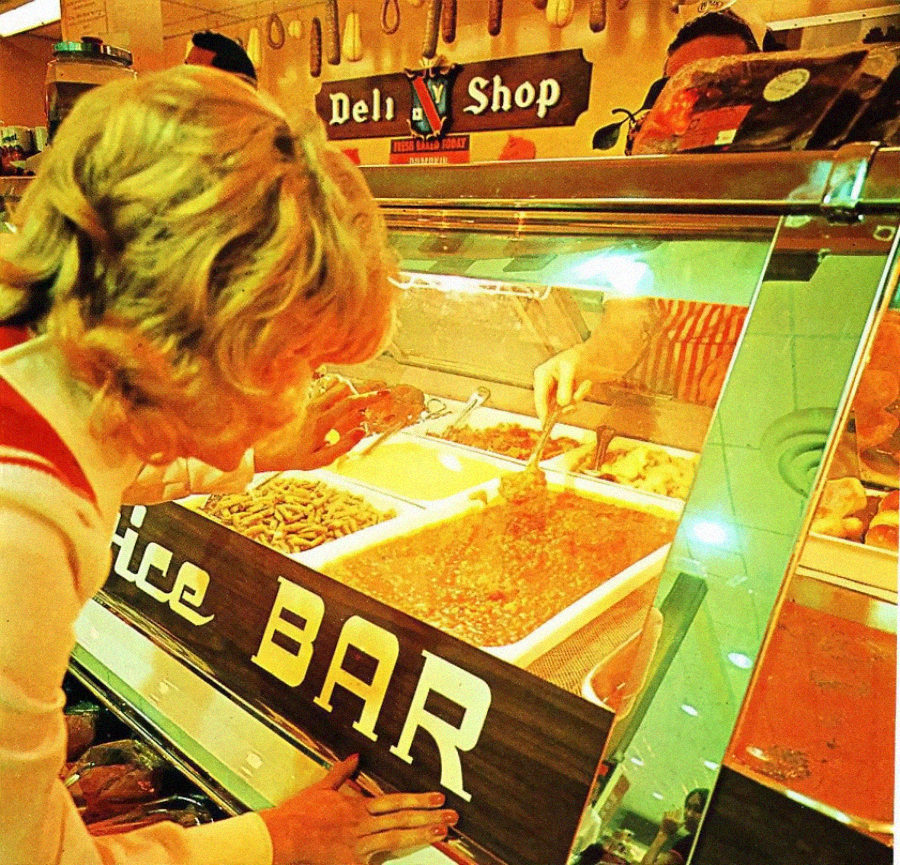 A person at a deli shop counter selects food from a hot food bar, while an employee behind the glass serves a dish with a ladle. The deli sign and assorted foods are visible in a warm, yellow-toned light.