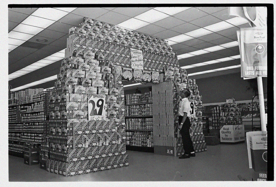 A black-and-white photo shows a grocery store display made of stacked cans forming an archway. A large sign reads "SPECIAL 29¢." A person in uniform stands inside the arch, surrounded by store shelves.