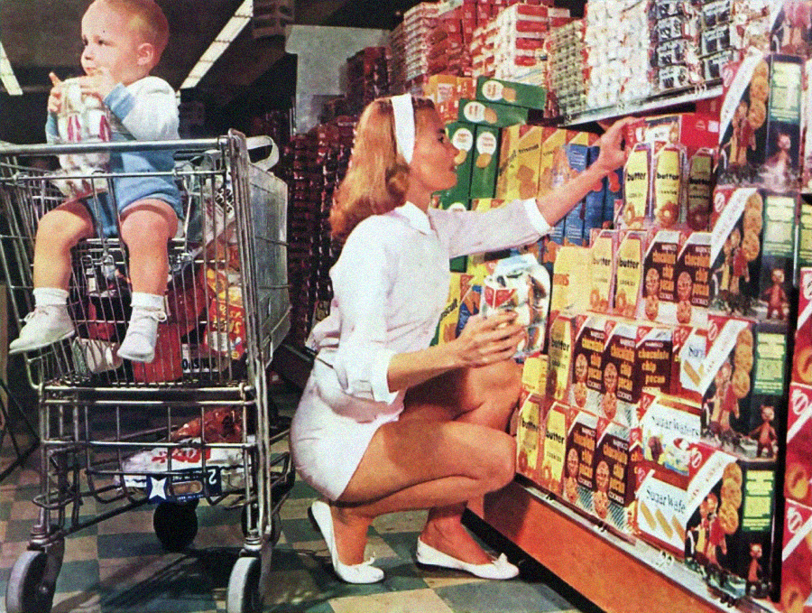 A woman in a white outfit kneels in a grocery store aisle, selecting packaged food from shelves. A young child sits in a shopping cart, holding a box and looking to the side. The aisle is lined with colorful boxes and cans.