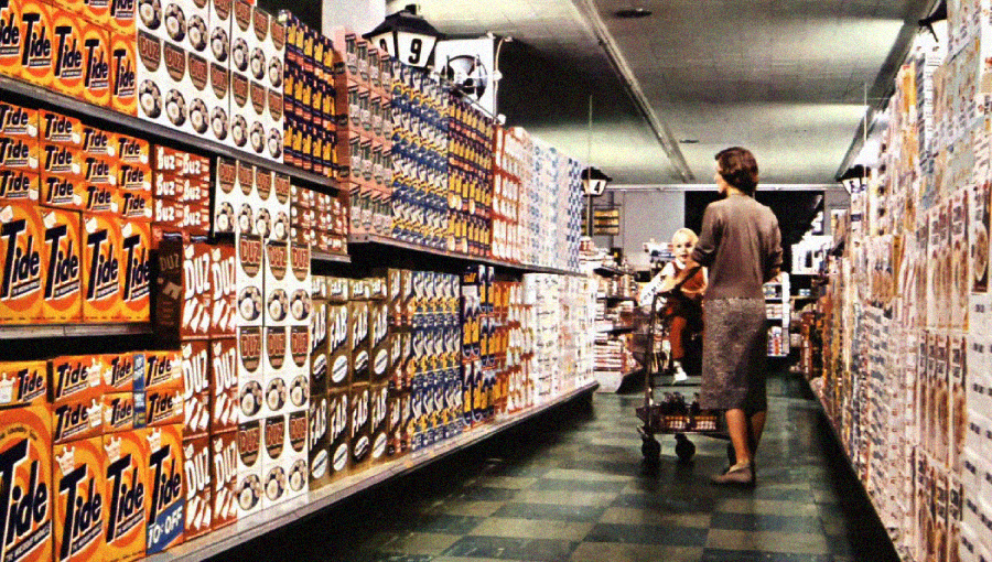 A woman pushes a shopping cart with a child in a grocery store aisle lined with shelves full of laundry detergent boxes, including Tide, under fluorescent lighting.