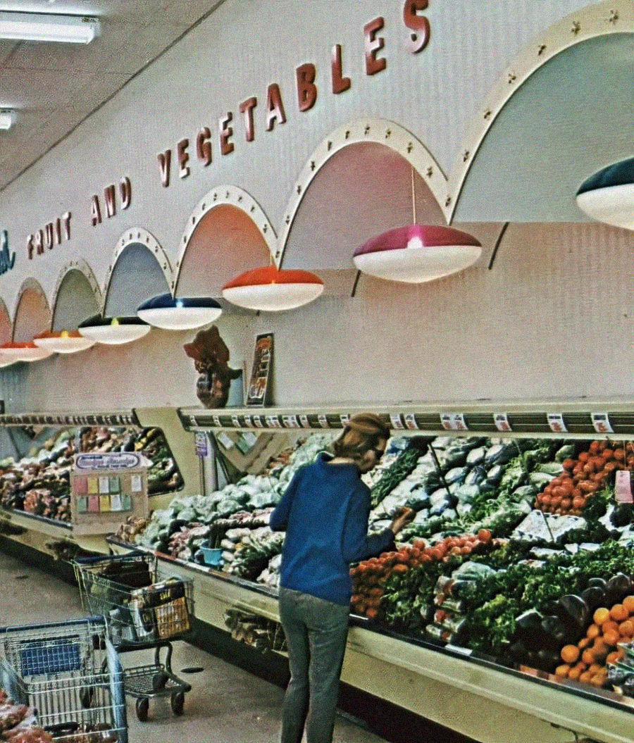 A person in a blue jacket selects produce from a brightly lit supermarket display. The section is labeled "Fruit and Vegetables" above colorful dome lights. Shopping carts and shelves of fresh produce are visible.