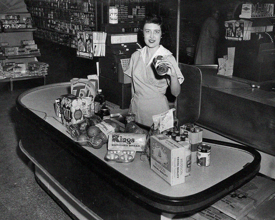 A woman in a uniform stands behind a vintage supermarket checkout counter, holding a can. Various groceries, including bread, canned goods, and produce, are on the counter. The store shelves are stocked in the background.