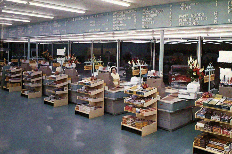 A vintage supermarket checkout area with several empty aisles, candy displays, a cashier in uniform, and store directory signs listing grocery items above the registers; cars are visible through the windows.